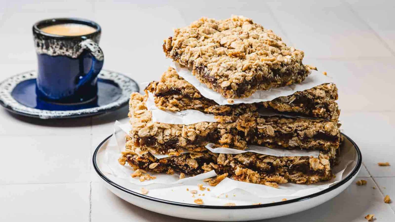 A stack of crumbly oat bars on a plate with parchment paper, next to a cup of coffee.