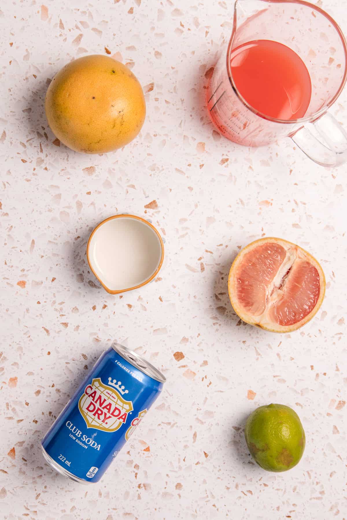 Grapefruit, lime, grapefruit juice, cream, and a can of Canada Dry Club Soda on a speckled countertop come together for a refreshing Sparkling Grapefruit Mocktail—an easy non-alcoholic drink for any occasion.