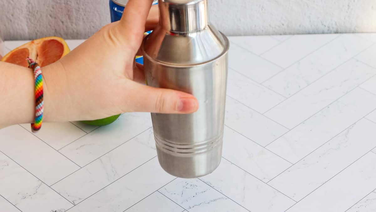 Hand holding a stainless steel cocktail shaker on a white tiled surface; ingredients for a refreshing grapefruit mocktail, including grapefruit and lime, are nearby.