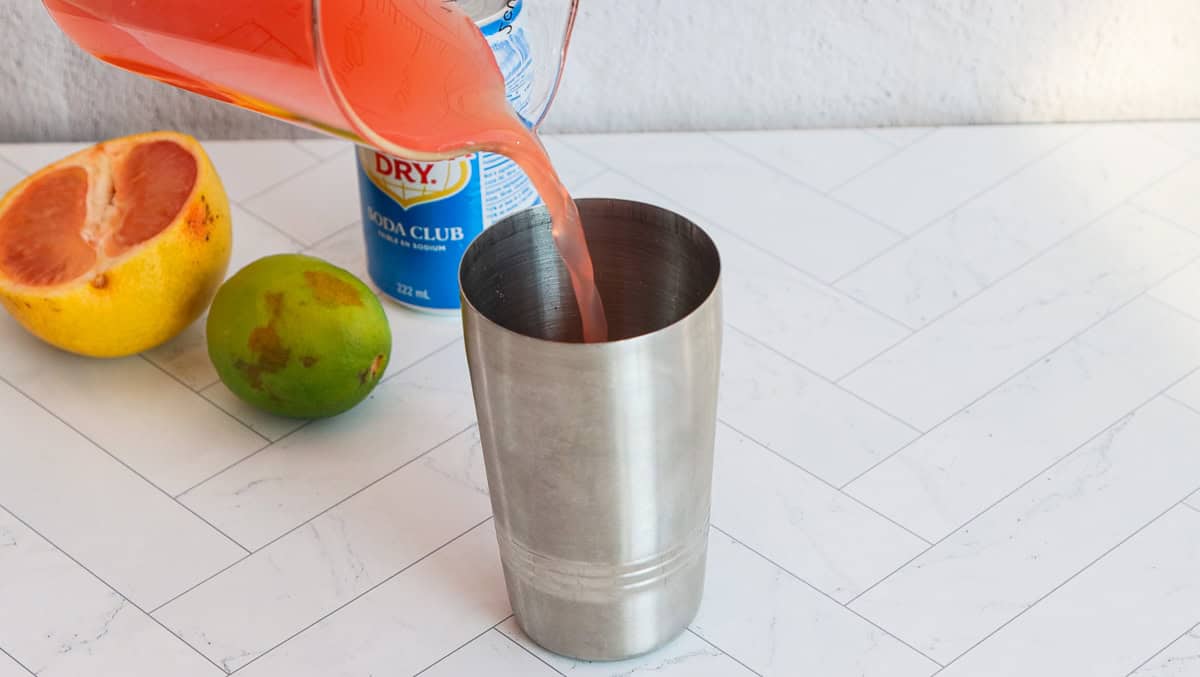 Grapefruit juice is poured into a metal shaker with fresh grapefruit, lime, and soda can on a white counter, ready to craft a refreshing grapefruit mocktail.