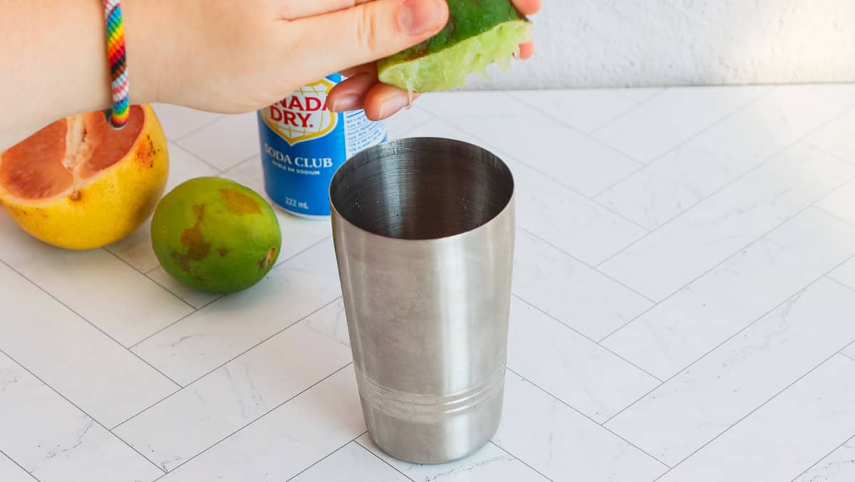 A hand squeezes a lime over a metal cocktail shaker on a white counter, preparing a refreshing grapefruit mocktail with fruit and soda nearby.