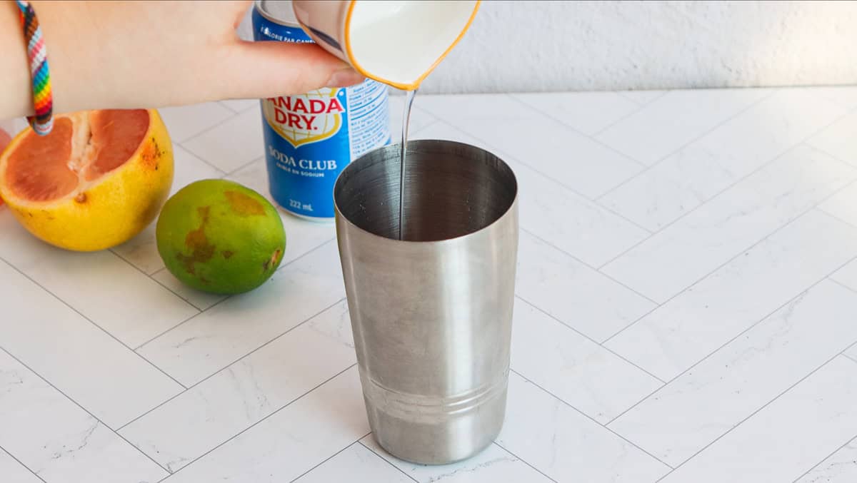 A hand pours liquid into a metal shaker; a grapefruit, lime, and soda can sit on a white countertop, ready for crafting a refreshing grapefruit mocktail.