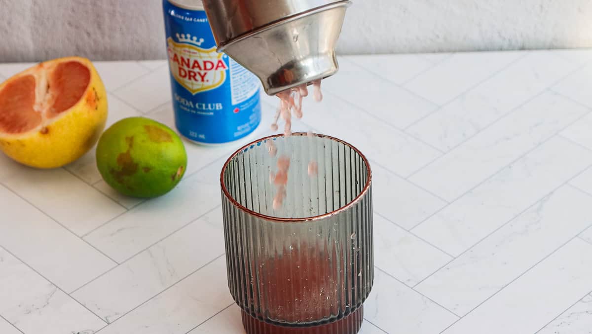A cocktail shaker pours pink liquid into a glass; grapefruit, lime, and a soda can are nearby on the counter, perfect for crafting a refreshing grapefruit mocktail.