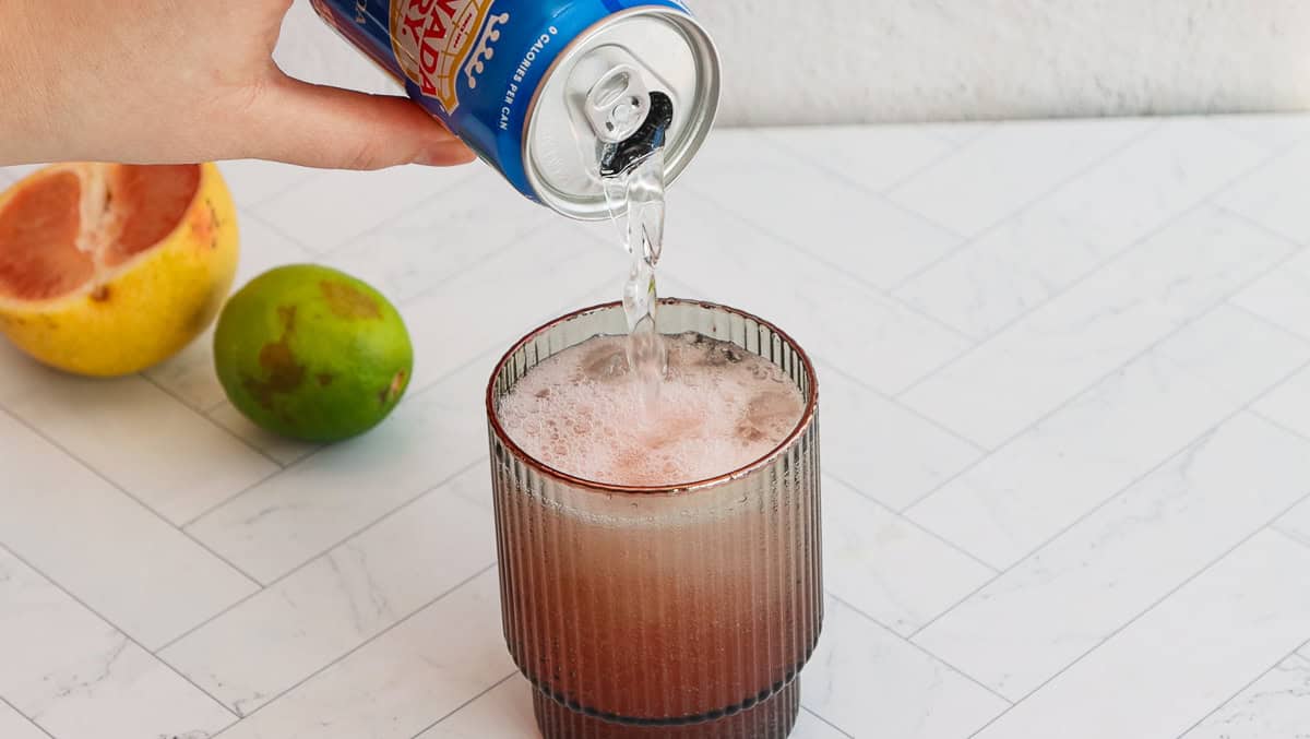A hand pours sparkling water from a can into a glass with a pink grapefruit mocktail, near a lime and grapefruit.