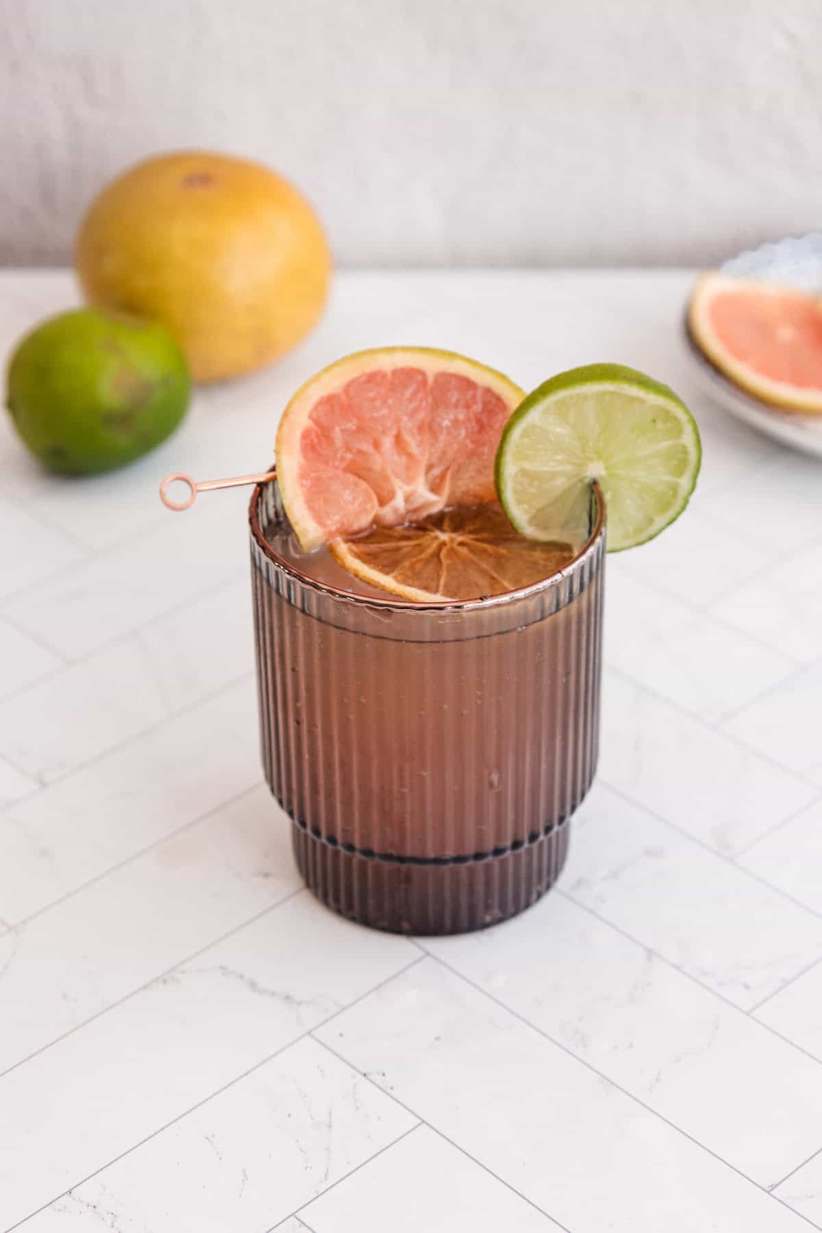 A brown glass filled with a Sparkling Grapefruit Mocktail, garnished with grapefruit and lime slices, sits on a white surface with fresh citrus fruits in the background.