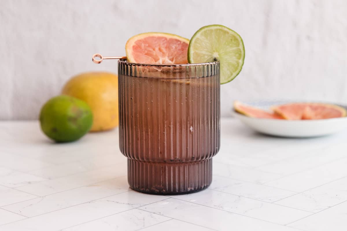 A sparkling grapefruit mocktail garnished with grapefruit and lime slices in a ribbed glass, with fresh fruit in the background.