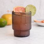 A dark glass with a Sparkling Grapefruit Mocktail, garnished with grapefruit and lime slices, sits on a white surface.
