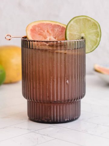 A dark glass with a Sparkling Grapefruit Mocktail, garnished with grapefruit and lime slices, sits on a white surface.
