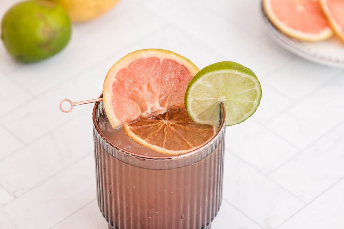 A sparkling mocktail in a ribbed glass, garnished with slices of grapefruit and lime, sits atop a white tiled surface.