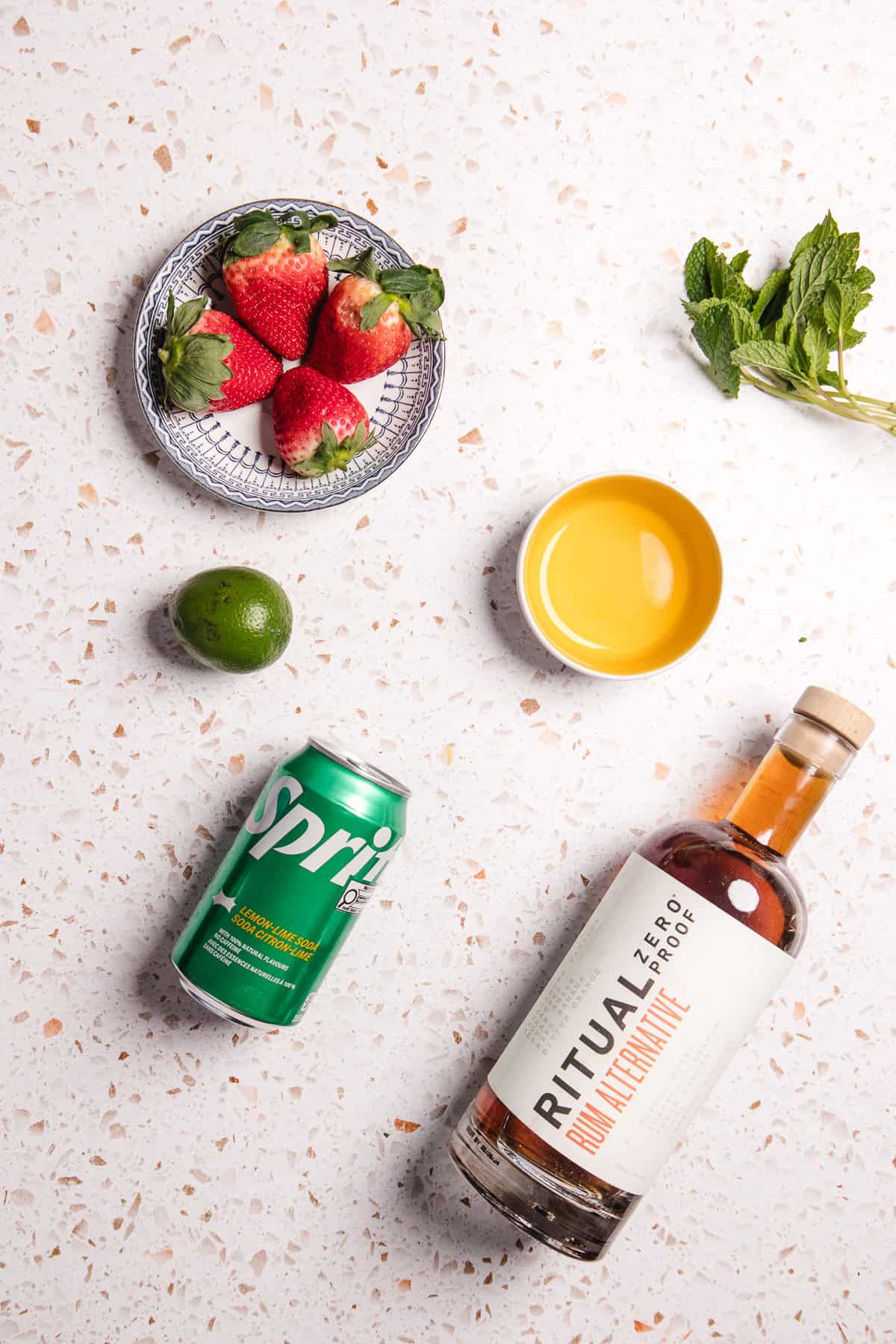 A bottle of Ritual rum alternative, a can of Sprite, strawberries, lime, mint, and a bowl of yellow liquid ready to create a refreshing strawberry lime mojito mocktail.