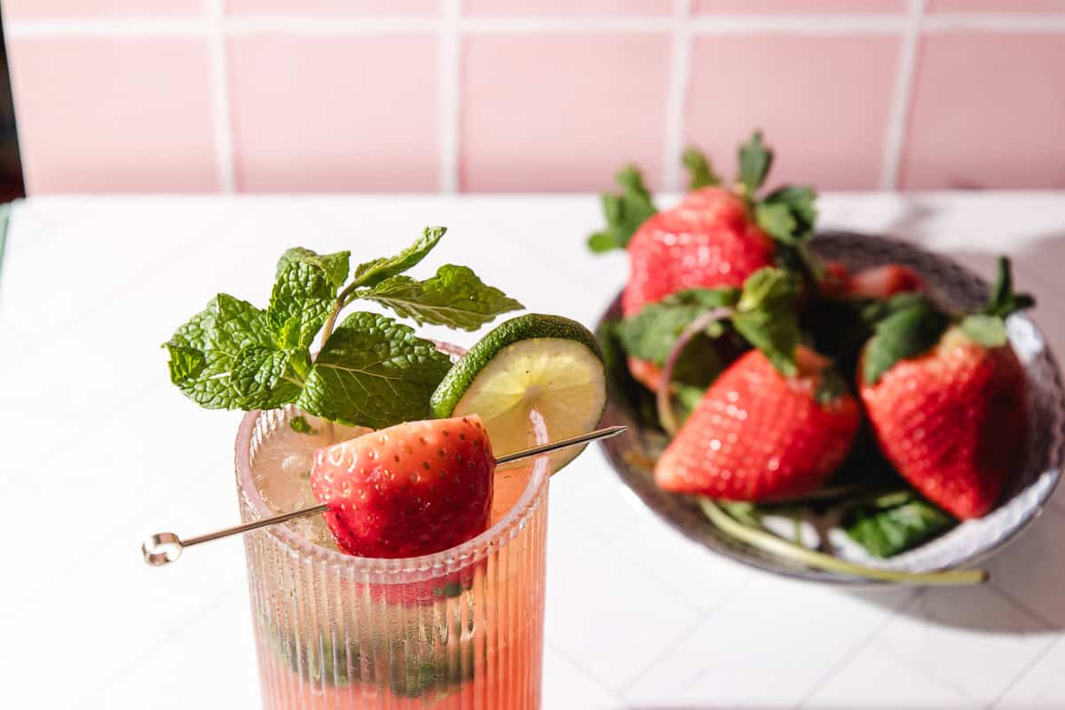 A strawberry lime mojito mocktail garnished with mint, lime, and a strawberry, served alongside a bowl of fresh strawberries.