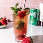 A tall glass of strawberry lime mojito mocktail with mint, lime, strawberry, and a striped straw, served over ice and a Sprite can in the background.