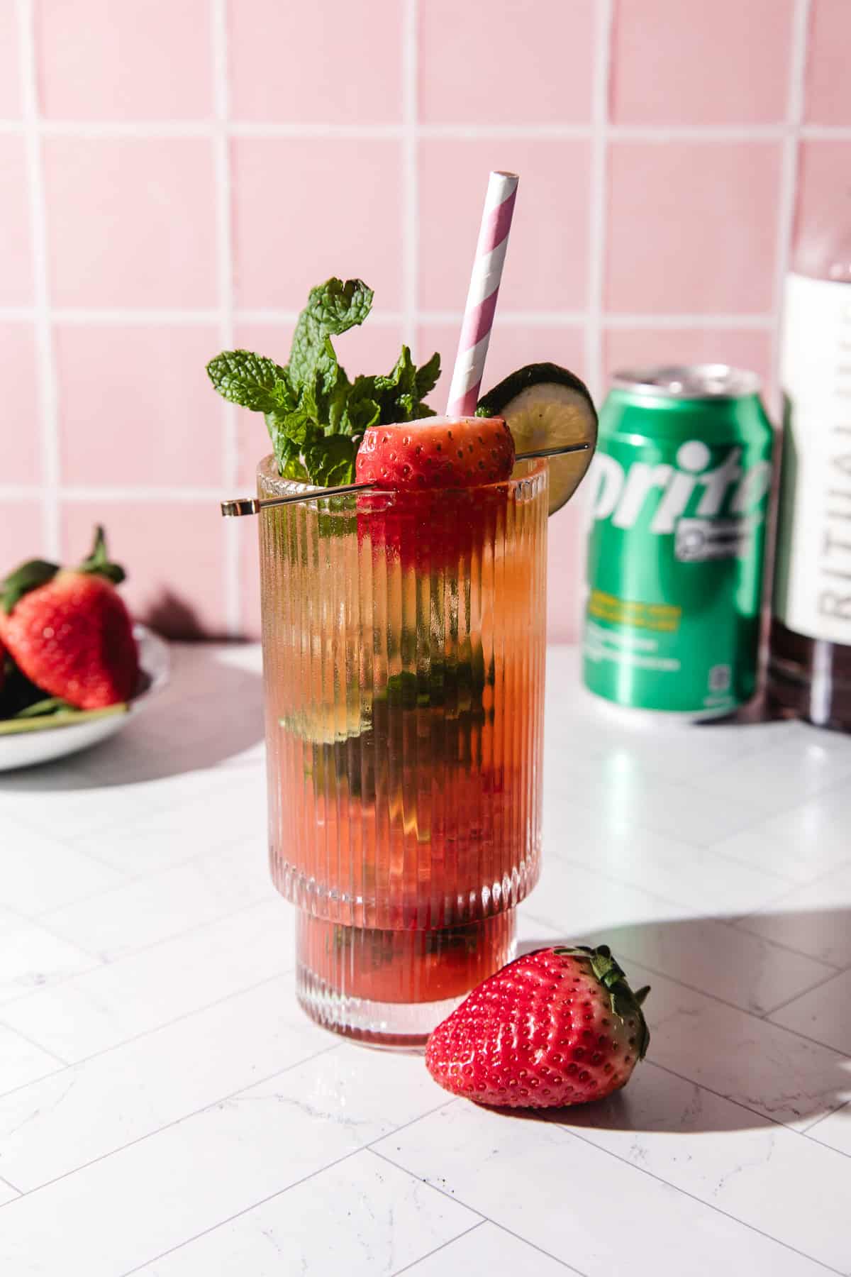A tall glass of strawberry lime mojito mocktail with mint, lime, strawberry, and a striped straw, served over ice and a Sprite can in the background.