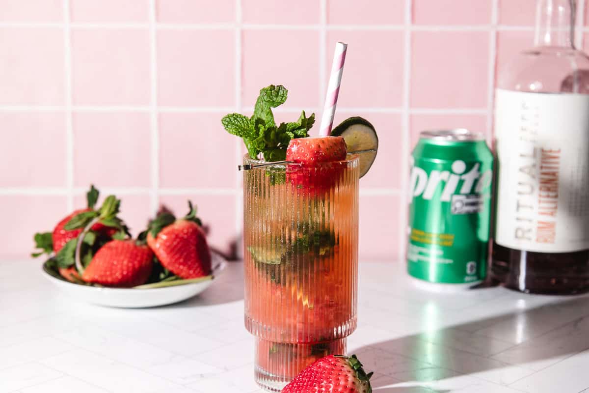 A strawberry lime mojito mocktail in a glass with a straw, surrounded by fresh strawberries and a can of Sprite.