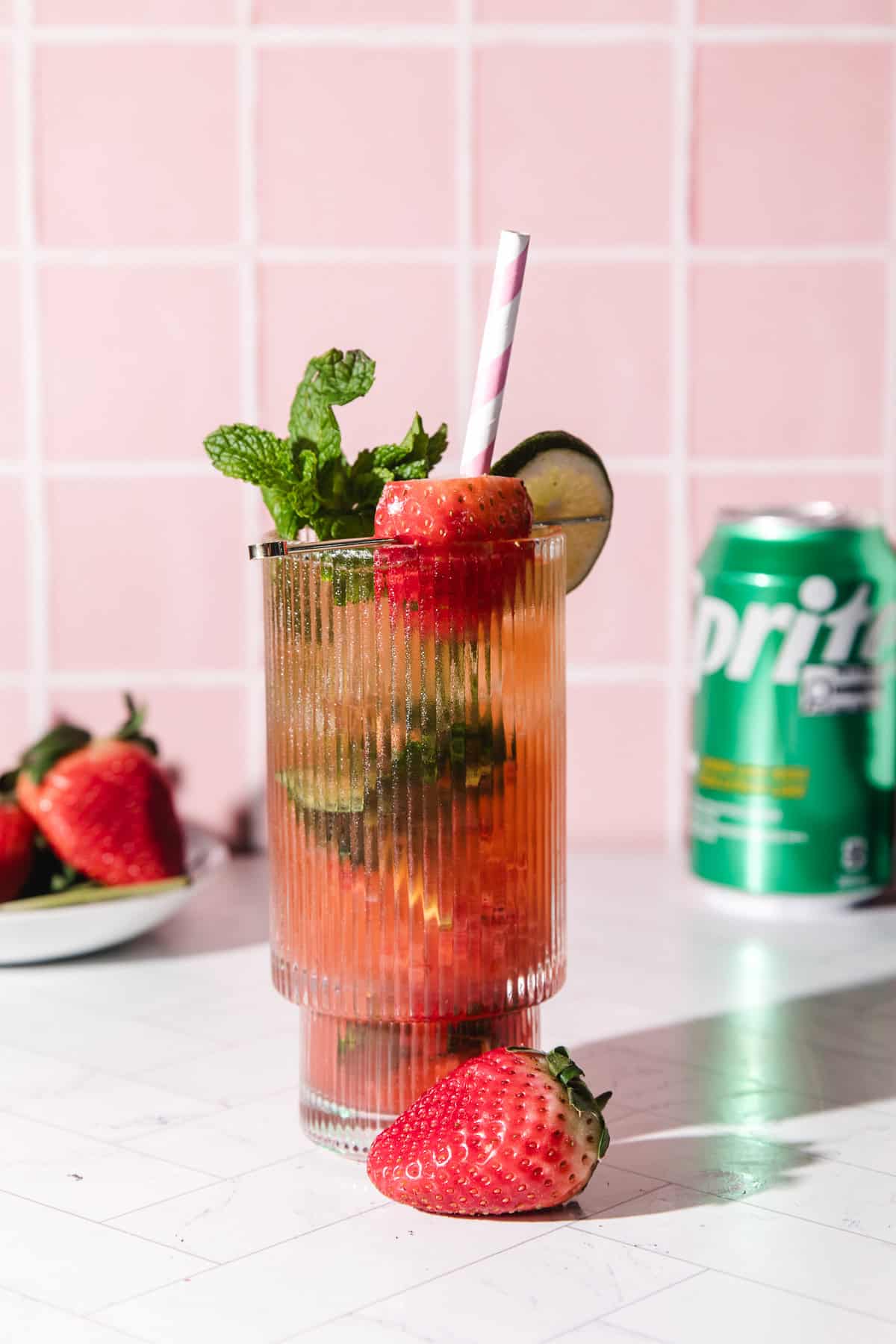 A strawberry lime mojito mocktail with mint, lime, and a straw sits beside fresh strawberries and a can of Sprite.