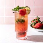 A strawberry lime mojito mocktail in a tall glass with mint and a lime slice, served beside a bowl of fresh strawberries.