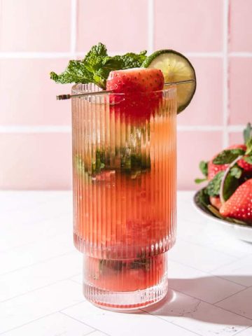 A strawberry lime mojito mocktail in a tall glass with mint and a lime slice, served beside a bowl of fresh strawberries.