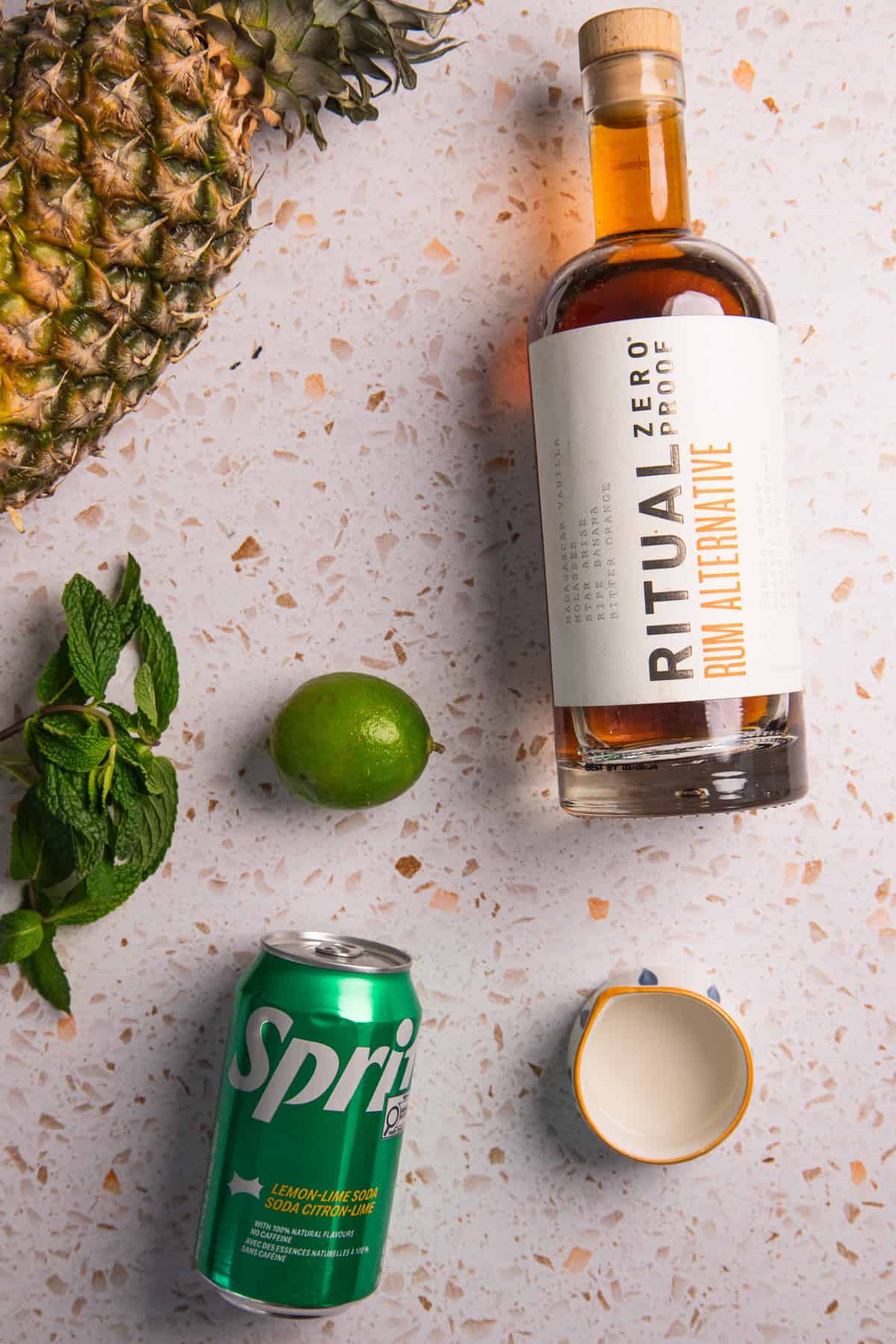 Flat lay of ingredients for a pineapple mojito mocktail: pineapple, mint, lime, Sprite can, a bottle of Ritual Zero Proof rum, and an empty cup on a counter.