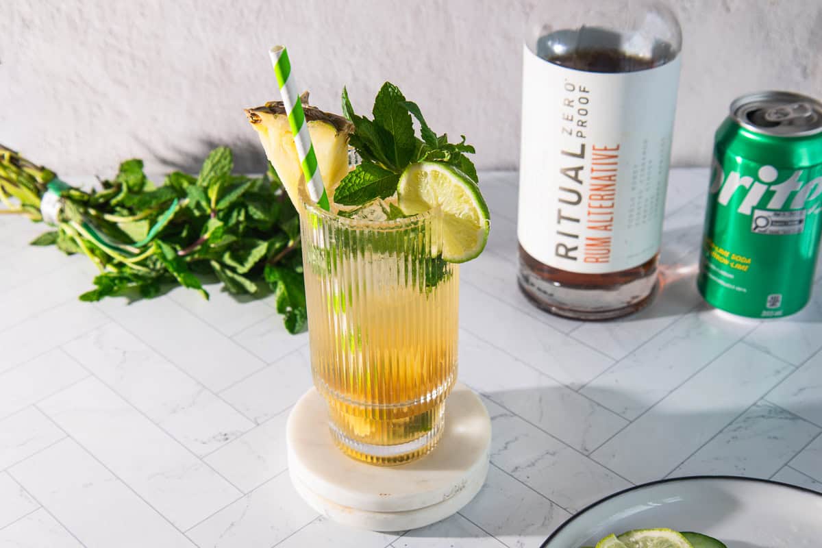 A pineapple mojito mocktail with lime and mint sits beside a bottle, soda can, and fresh mint on a marble surface.
