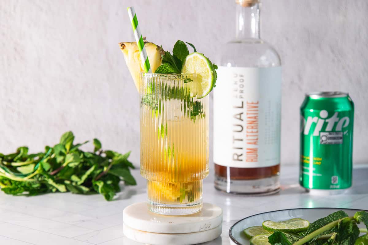 Tall glass of pineapple mojito mocktail garnished with lime, mint, and pineapple; bottle and can of Sprite in the background.