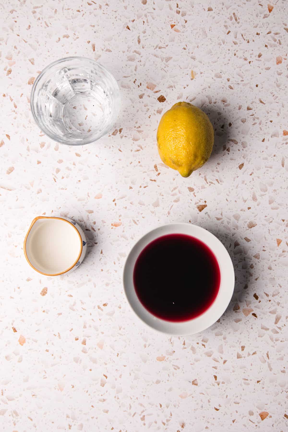 A glass of water, a whole lemon, a small cream pitcher, and a bowl of pomegranate lemonade sit on a speckled surface.
