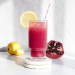 A glass of vibrant pomegranate lemonade with a lemon slice and straw sits beside fresh lemons and a pomegranate on a white surface.