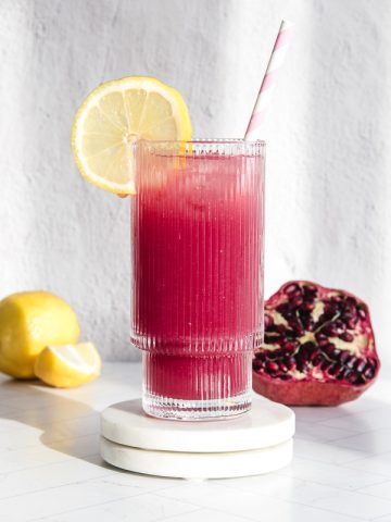 A glass of vibrant pomegranate lemonade with a lemon slice and straw sits beside fresh lemons and a pomegranate on a white surface.