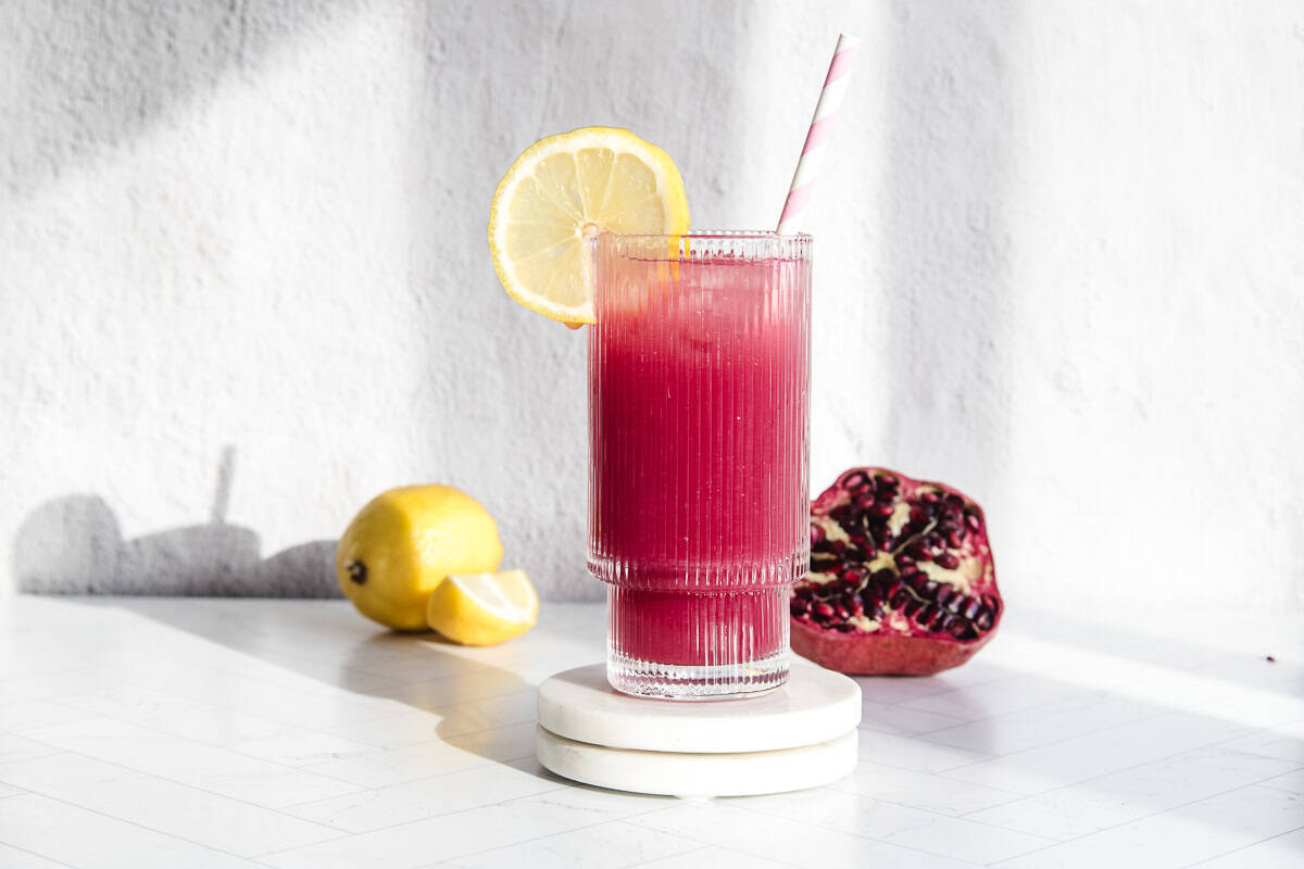 A glass of pomegranate lemonade with a lemon slice and straw sits next to fresh lemons and a pomegranate on a white surface.