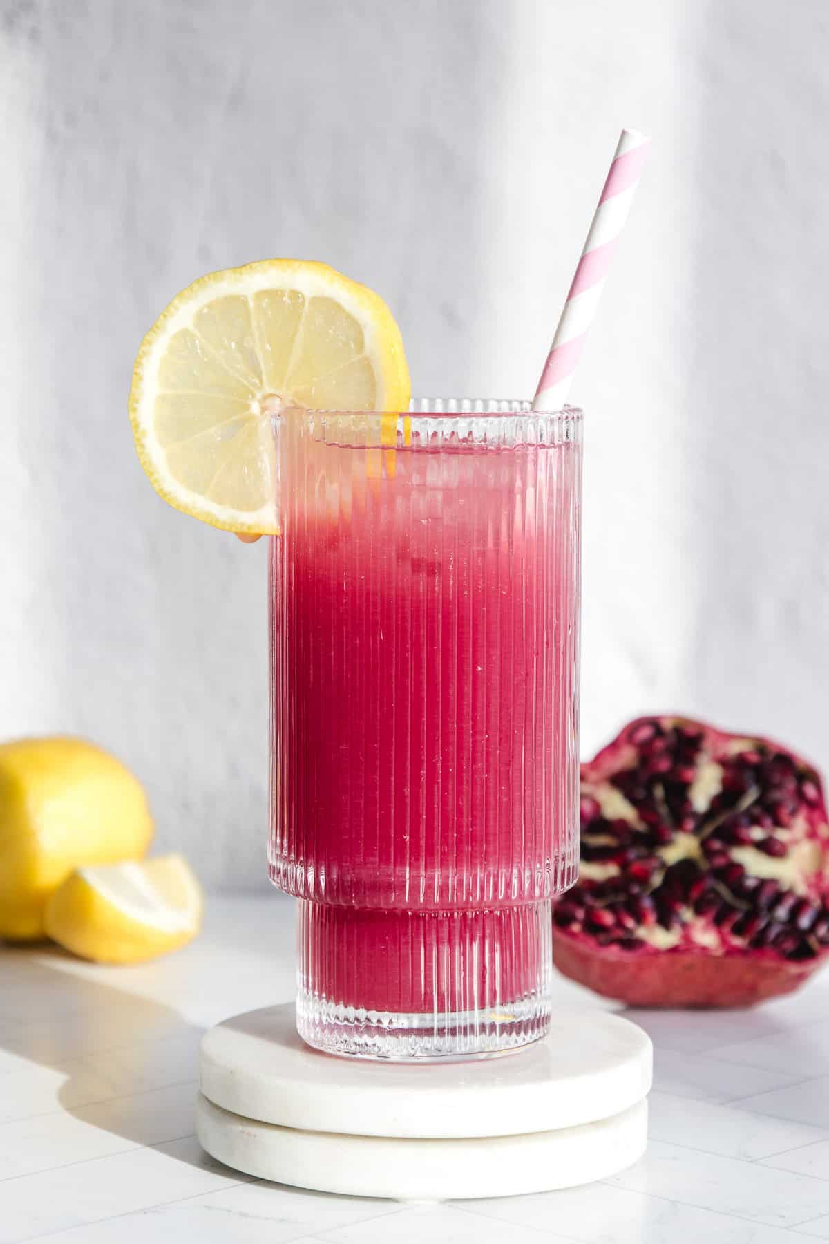 A glass of pink pomegranate lemonade with a lemon slice and striped straw, surrounded by fresh lemons and pomegranate in the background.
