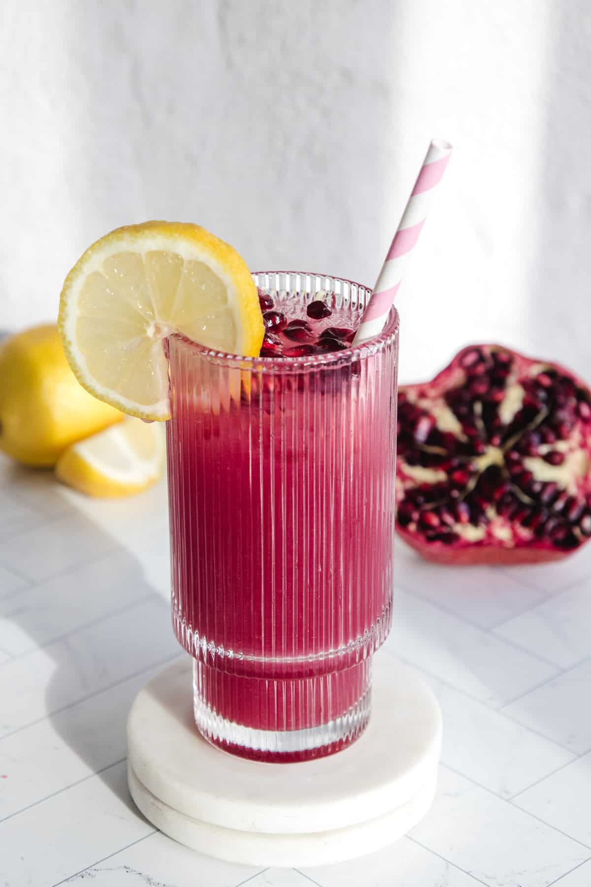 A glass of pomegranate lemonade with a lemon slice, pink straw, and fresh pomegranates and lemons in the background.