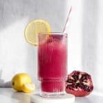 A pink pomegranate lemonade in a glass with a lemon slice, striped straw, and fresh lemon and pomegranate in the background.