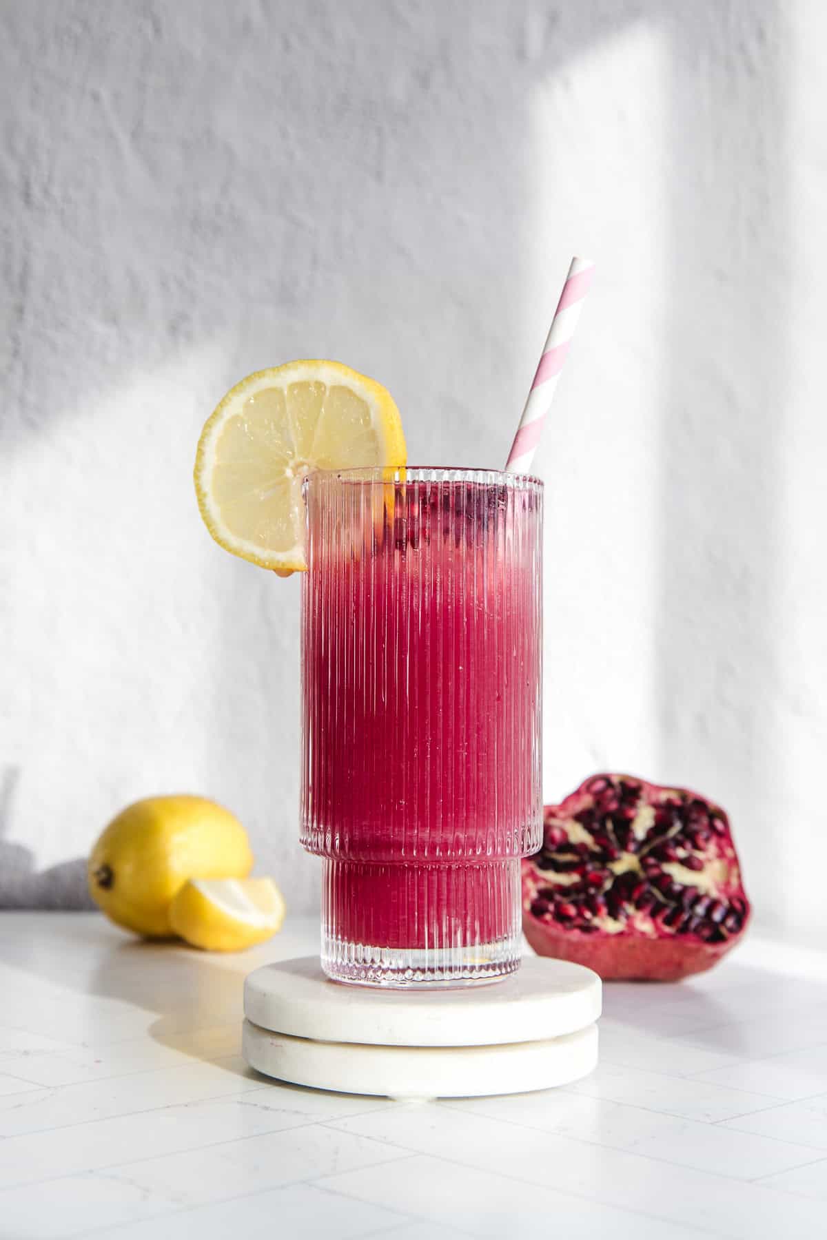 A pink pomegranate lemonade in a glass with a lemon slice, striped straw, and fresh lemon and pomegranate in the background.