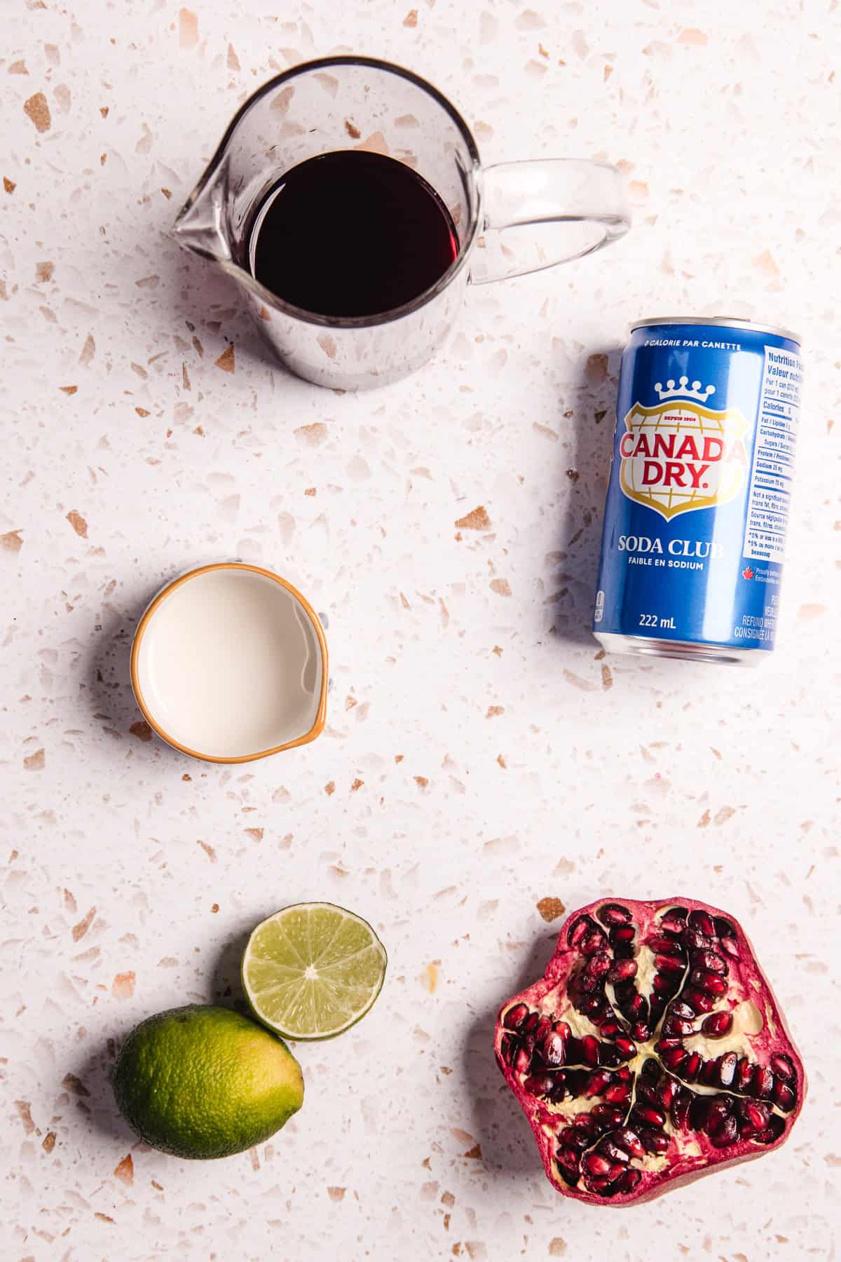 Ingredients on a countertop: pomegranate, lime, cream, dark liquid in a pitcher, and a can of Canada Dry soda&mdash;perfect for crafting a refreshing pomegranate mocktail.
