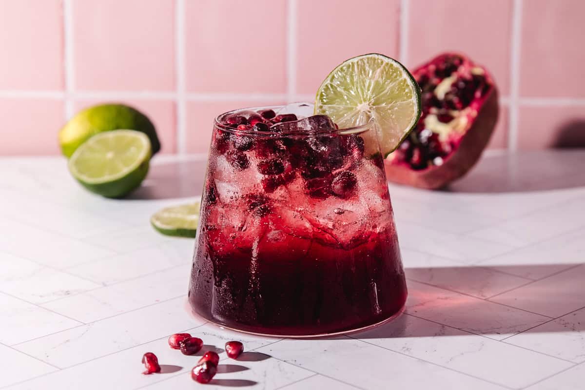 A glass of refreshing pomegranate mocktail with ice, garnished with a lime slice, sits on a white countertop.