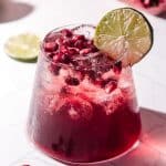 A glass of refreshing pomegranate mocktail with ice, pomegranate seeds, and a lime slice garnish on a white surface.