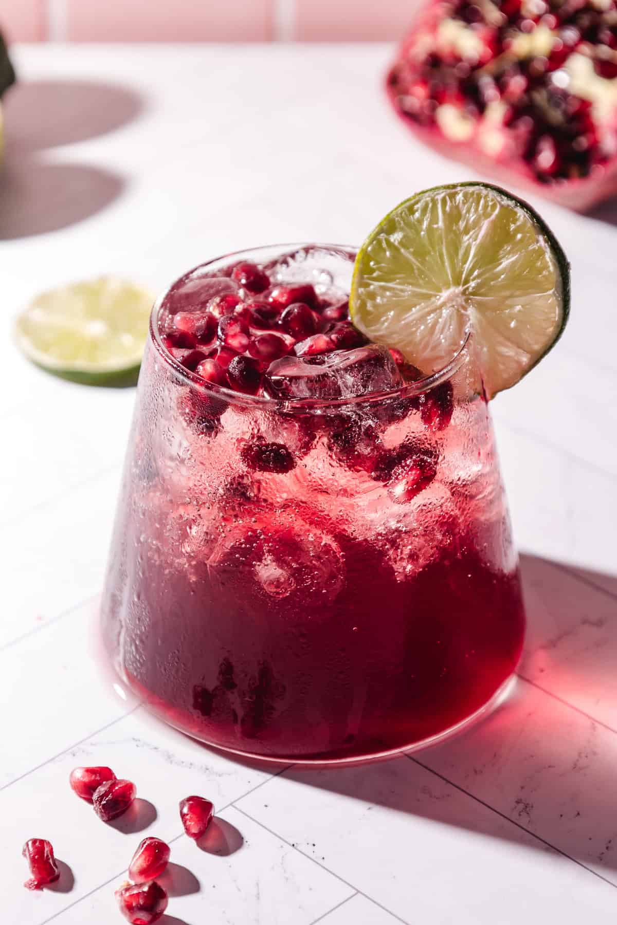 A glass of refreshing pomegranate mocktail with ice, pomegranate seeds, and a lime slice garnish on a white surface.