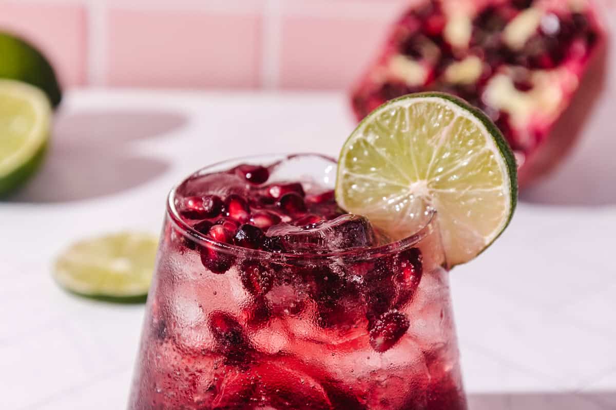 A refreshing pomegranate mocktail served over ice in a glass, garnished with a lime slice, with fresh pomegranate and limes in the background.