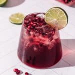 A refreshing glass of pomegranate mocktail, garnished with pomegranate seeds and a lime slice, served on a white surface-a perfect non-alcoholic drink for any occasion.