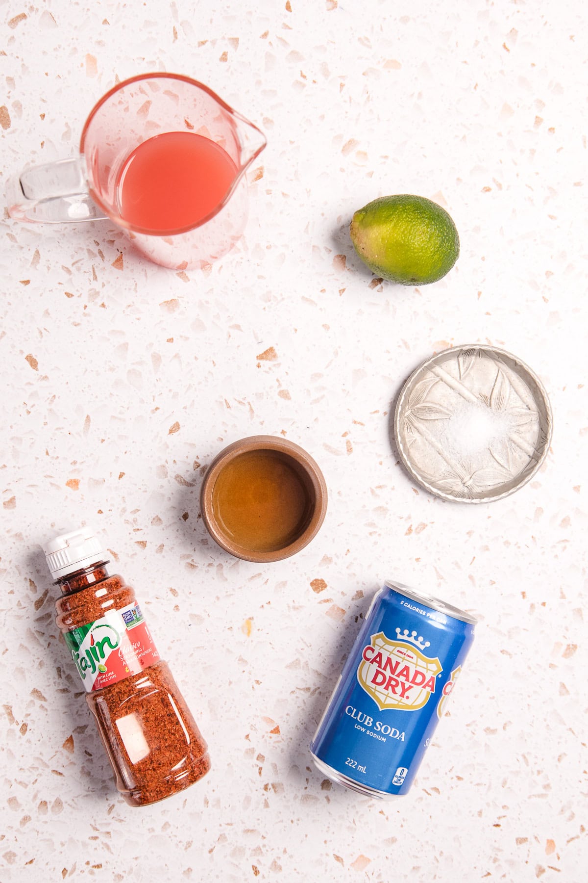 Ingredients for a Spicy Grapefruit Margarita Mocktail: lime, Taj&iacute;n, grapefruit juice, simple syrup, sugar, and club soda on a counter for a refreshing non alcoholic treat.