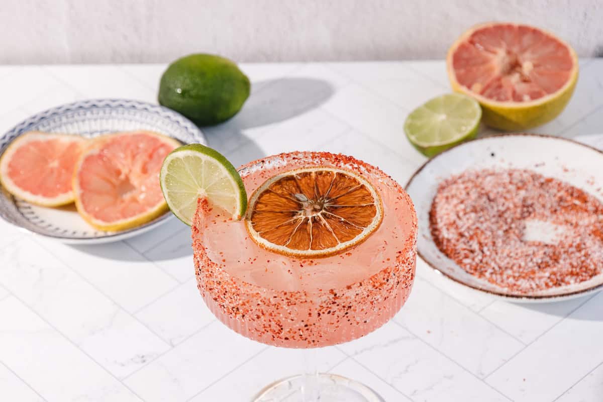 A pink Margarita Mocktail with a chili salt rim, garnished with lime and dried citrus, sits near grapefruit and lime slices&mdash;this non-alcoholic drink offers a refreshing, spicy grapefruit twist.