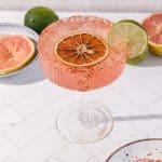 A spicy grapefruit mocktail in a glass rimmed with chili, garnished with dried orange and lime slices, sits on a white table.