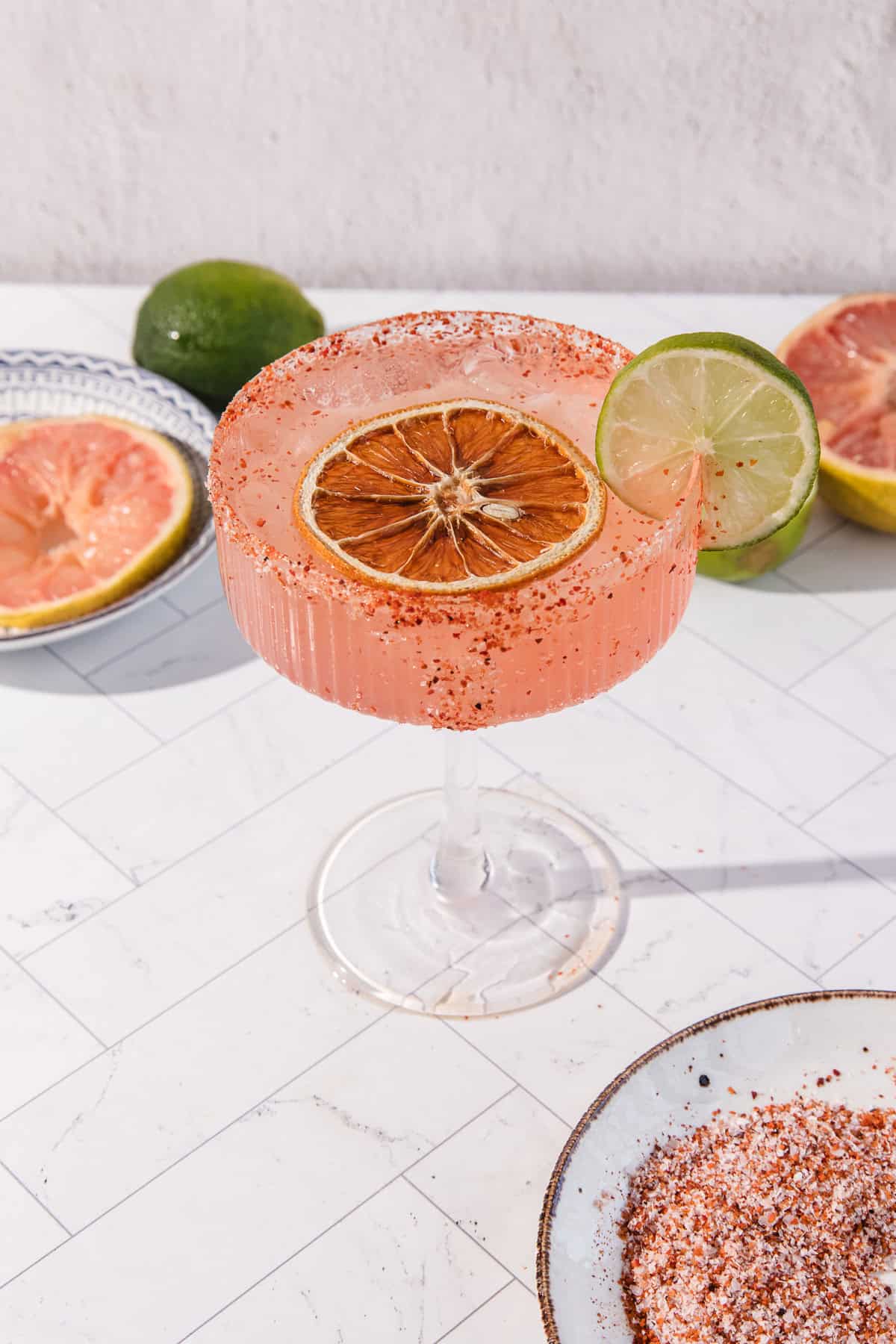 A spicy grapefruit mocktail in a glass rimmed with chili, garnished with dried orange and lime slices, sits on a white table.