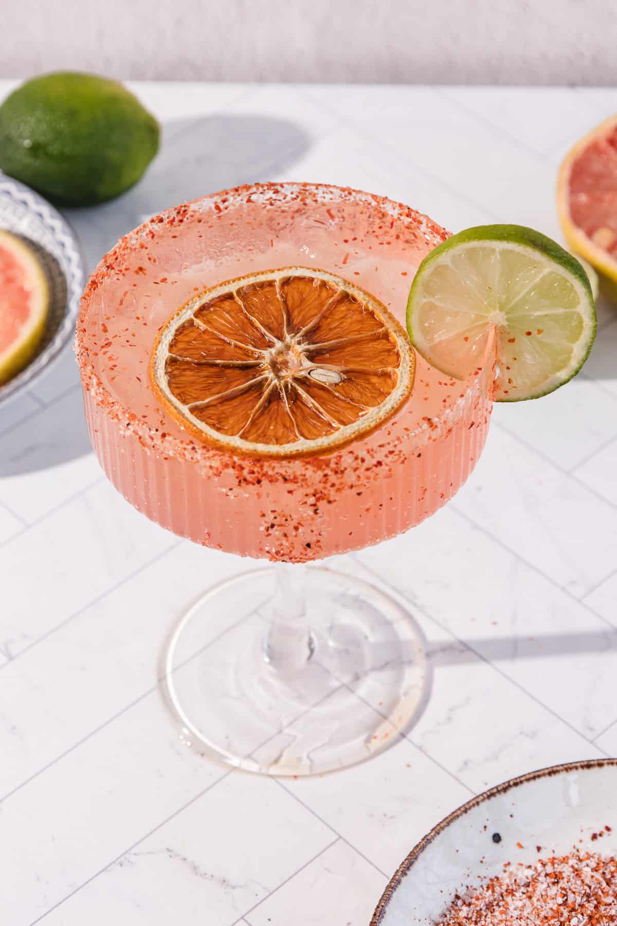 A pink Spicy Grapefruit Margarita Mocktail in a glass with a chili rim, garnished with dried orange and lime slice.