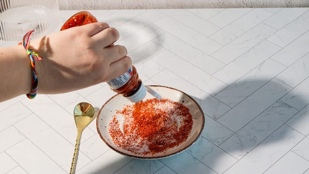 A hand pours hot sauce over a plate of red chili powder and salt, perfect for rimming a glass for a spicy margarita mocktail, with a spoon nearby on a tiled surface.
