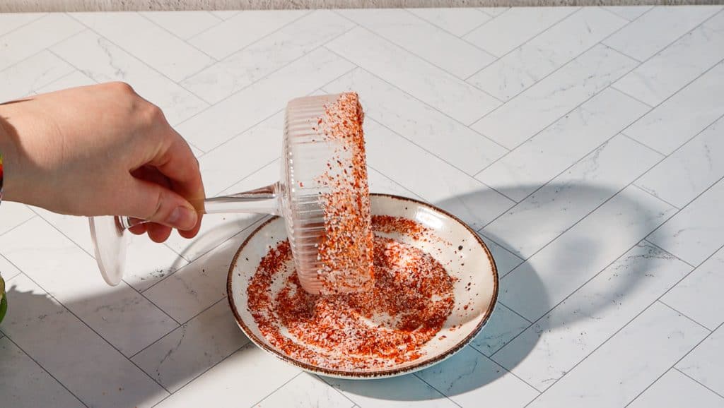 A hand dips a glass rim into a plate of red and white seasoning on a white tiled surface, prepping it for a spicy margarita mocktail.