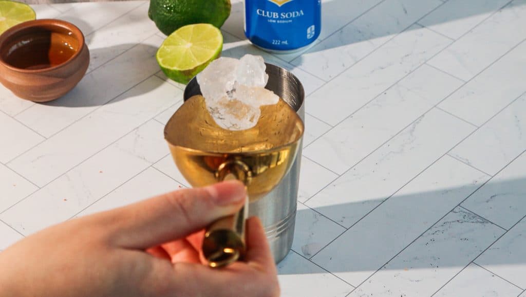 A hand holds a gold jigger above a shaker with ice; ingredients for a spicy margarita mocktail&mdash;lime, soda, and a bowl&mdash;rest on a white countertop.