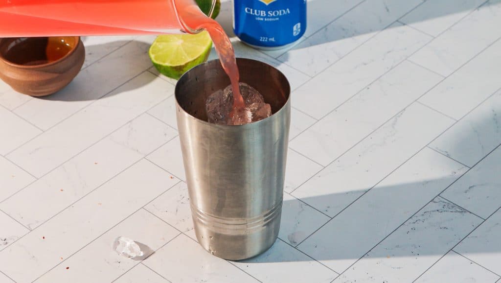 A spicy margarita mocktail is being poured into a metal cocktail shaker with ice, on a white tiled surface with lime and soda nearby.