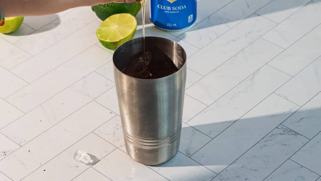 A hand stirs a spicy margarita mocktail with ice in a metal shaker on a tile countertop, with lime and club soda nearby.