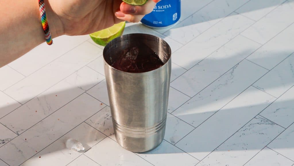 A hand squeezes a lime over a metal cocktail shaker filled with ice on a tiled surface, preparing a spicy margarita mocktail.