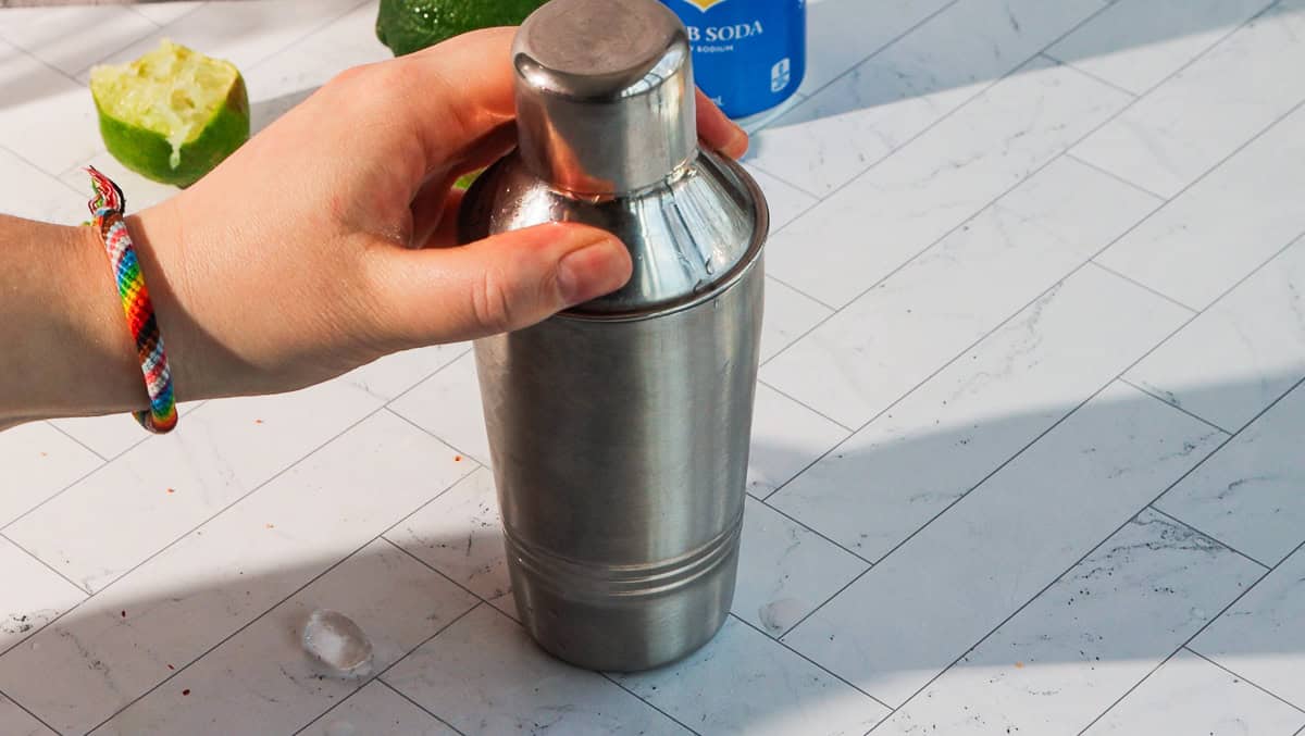 A hand holding a stainless steel cocktail shaker on a white tiled surface with a lime and soda in the background, ready to craft a spicy margarita mocktail.